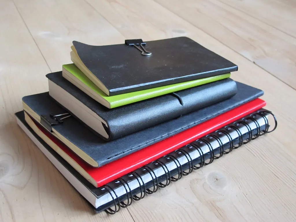 Stack of assorted notebooks with binder clips on a wooden floor